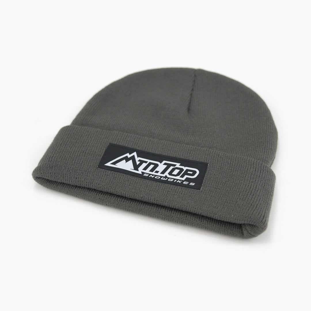 A dark gray knit beanie hat with a folded cuff, featuring a black rectangular patch on the front with the white text 'MTN.TOP Snowbikes' and a mountain graphic.
