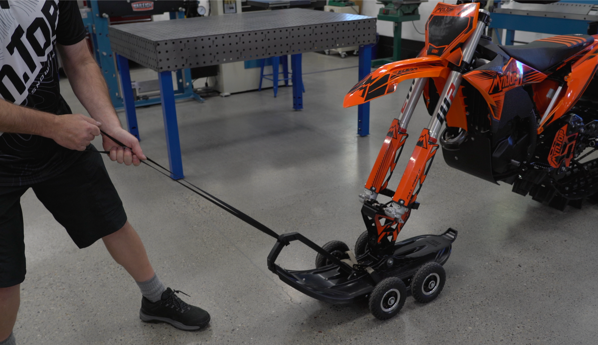 A person in the MTN.TOP shop pulls an orange KTM snowbike across the floor using a black MTN.TOP pull strap